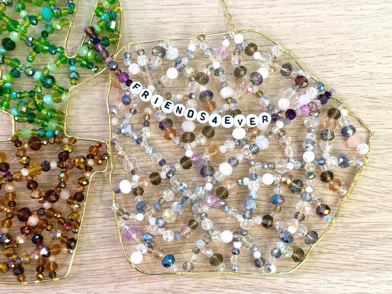 May include: Decorative beaded ornaments in the shape of a leaf and a geometric shape. The geometric shape has the text "FRIENDS4EVER" spelled out in white letter beads. The ornaments are made with gold wire and various colored beads.