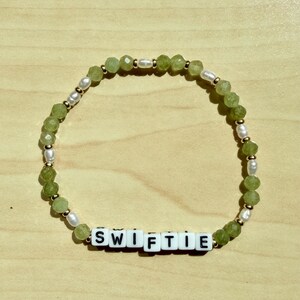 May include: A beaded bracelet with the word "SWIFTIE" spelled out in white letter beads. The bracelet features green faceted beads, small white pearls, and gold-colored spacers. The bracelet is displayed on a light brown wooden surface.