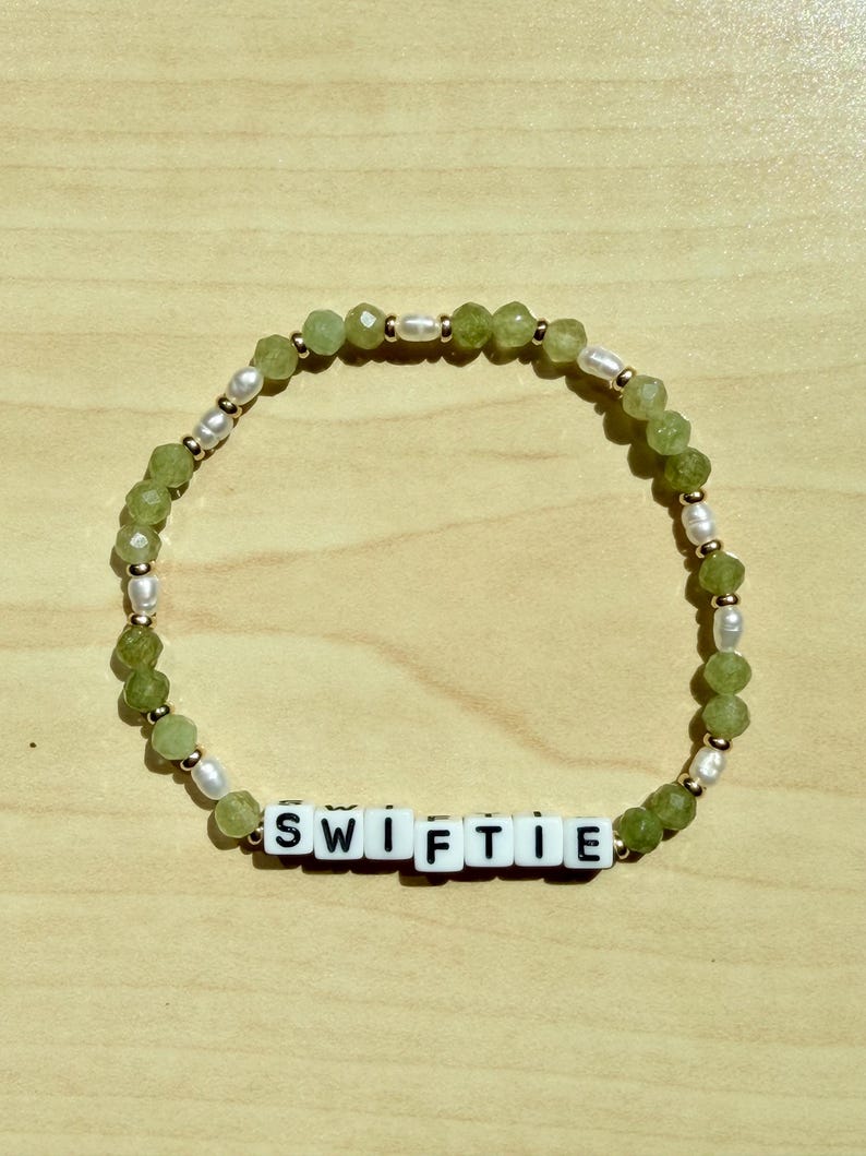 May include: A beaded bracelet with the word "SWIFTIE" spelled out in white letter beads. The bracelet features green faceted beads, small white pearls, and gold-colored spacers. The bracelet is displayed on a light brown wooden surface.