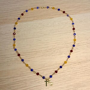 May include: A gold-toned chain necklace featuring a repeating pattern of small, faceted beads in blue, red, and yellow. The necklace includes two gold-toned charms hanging from the centre, one shaped like a cross and the other like the letter S.