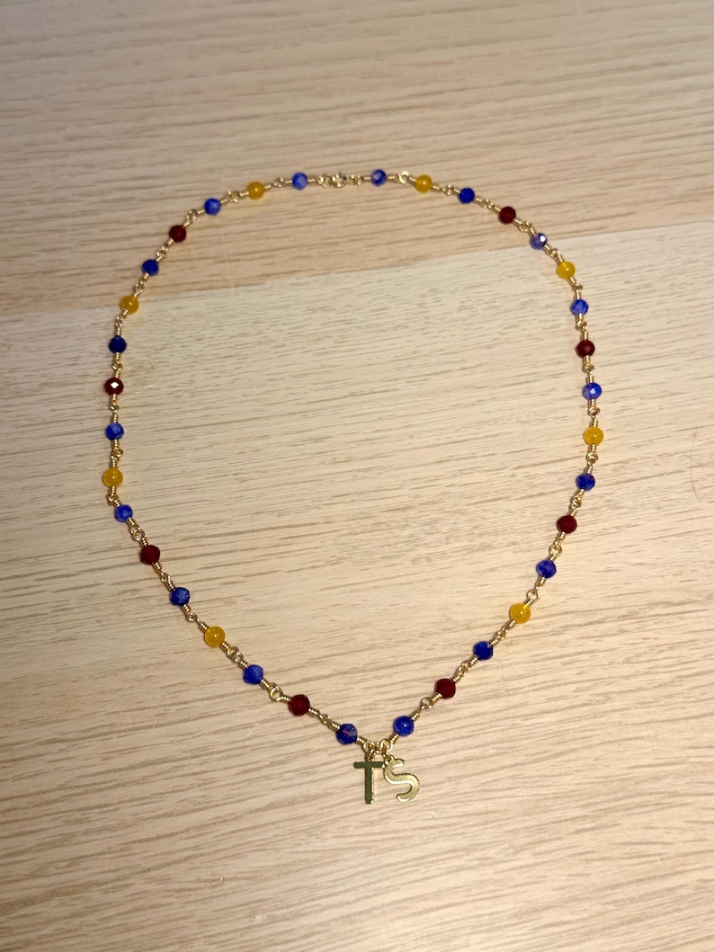 May include: A gold-toned chain necklace featuring a repeating pattern of small, faceted beads in blue, red, and yellow. The necklace includes two gold-toned charms hanging from the centre, one shaped like a cross and the other like the letter S.