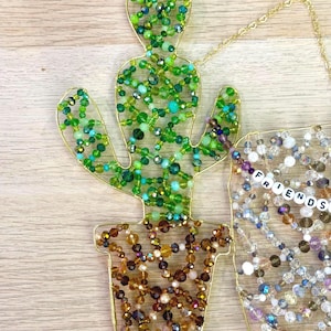 May include: A beaded cactus decoration with a gold wire frame. The cactus is filled with green beads, and the pot is filled with brown and gold beads. A separate beaded decoration spells out the word "FRIENDS".