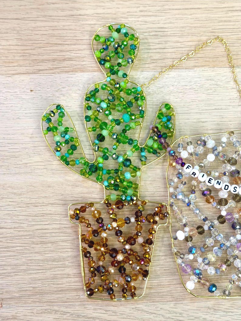May include: A beaded cactus decoration with a gold wire frame. The cactus is filled with green beads, and the pot is filled with brown and gold beads. A separate beaded decoration spells out the word "FRIENDS".
