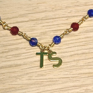 May include: A gold-toned chain necklace featuring two dangling letter charms, T and S, suspended from a wire-wrapped link design. The necklace is accented with small, faceted beads in alternating colours of deep red, royal blue, and dark blue.