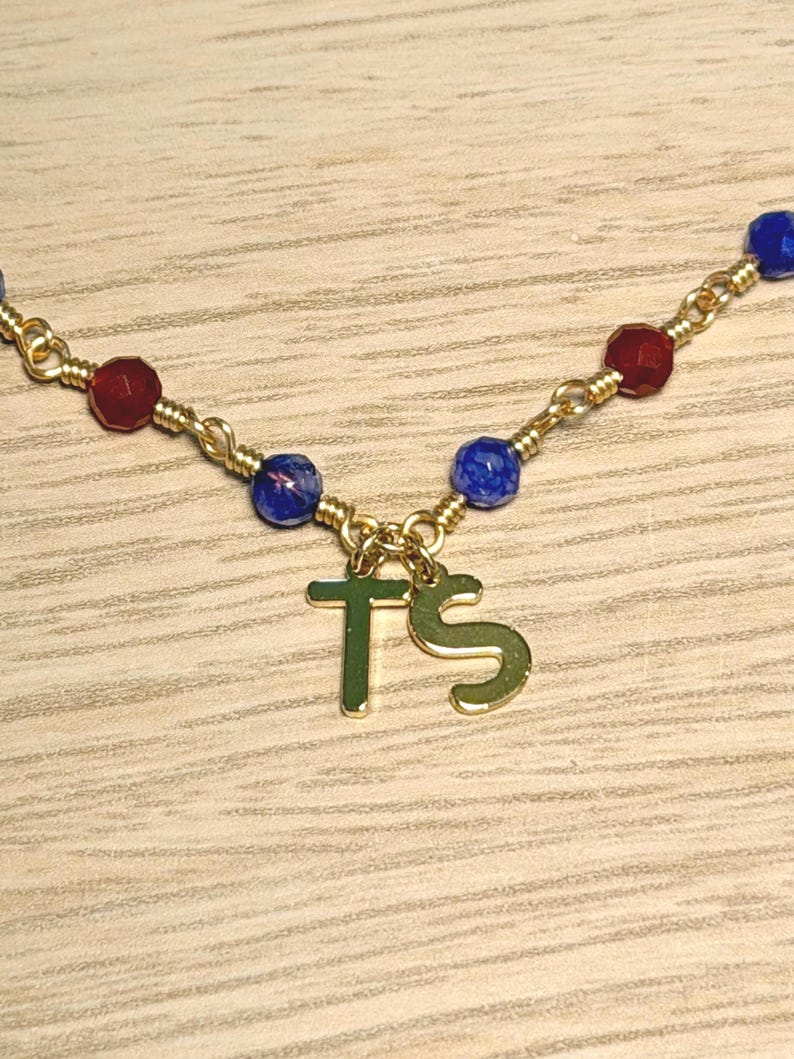 May include: A gold-toned chain necklace featuring two dangling letter charms, T and S, suspended from a wire-wrapped link design. The necklace is accented with small, faceted beads in alternating colours of deep red, royal blue, and dark blue.