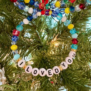 May include: A circular ornament made of multi-coloured glass and plastic beads in shades of red, yellow, blue, and light blue. The bottom section features white letter beads spelling out the phrase I LOVED YOU, separated by small gold-toned spacers.