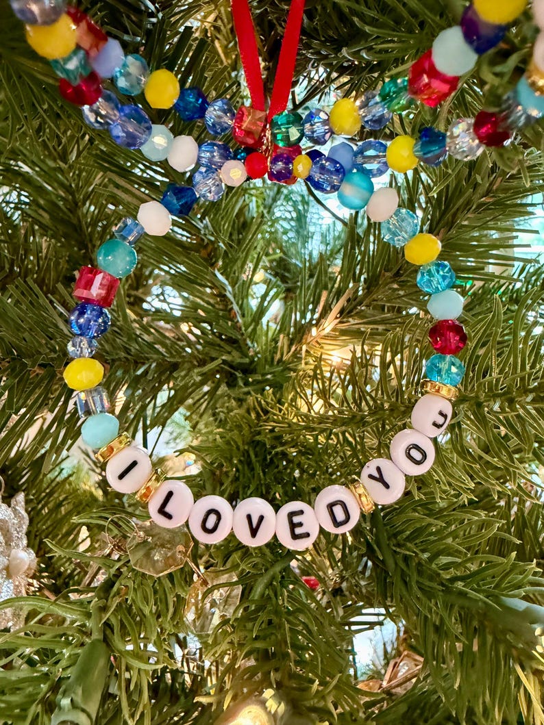 May include: A circular ornament made of multi-coloured glass and plastic beads in shades of red, yellow, blue, and light blue. The bottom section features white letter beads spelling out the phrase I LOVED YOU, separated by small gold-toned spacers.