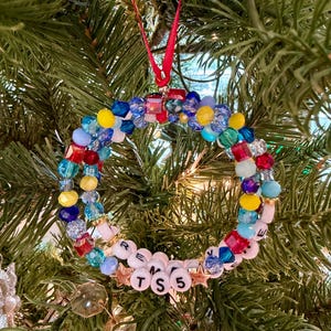 May include: A circular holiday ornament constructed from a variety of multi-coloured faceted beads in shades of red, blue, yellow, and light blue. The ornament features white letter beads spelling out TS 5 and is suspended by a thin red ribbon.