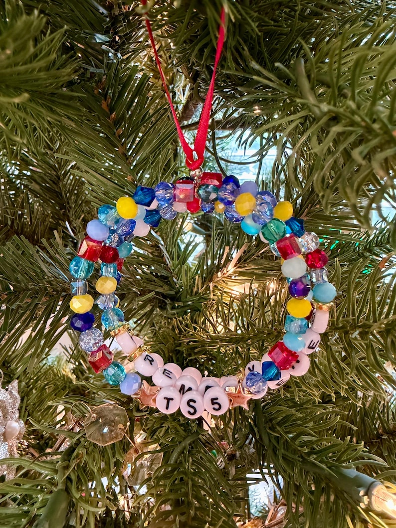 May include: A circular holiday ornament constructed from a variety of multi-coloured faceted beads in shades of red, blue, yellow, and light blue. The ornament features white letter beads spelling out TS 5 and is suspended by a thin red ribbon.