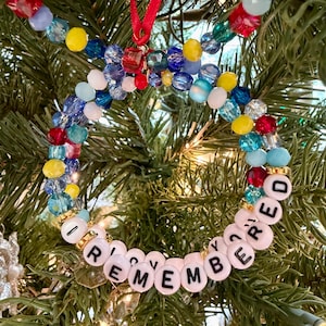 May include: A circular ornament made of multi-coloured faceted beads in red, blue, yellow, and clear tones. The bottom section features white round beads with black letters spelling out I REMEMBERED. A red ribbon loop is attached to the top.