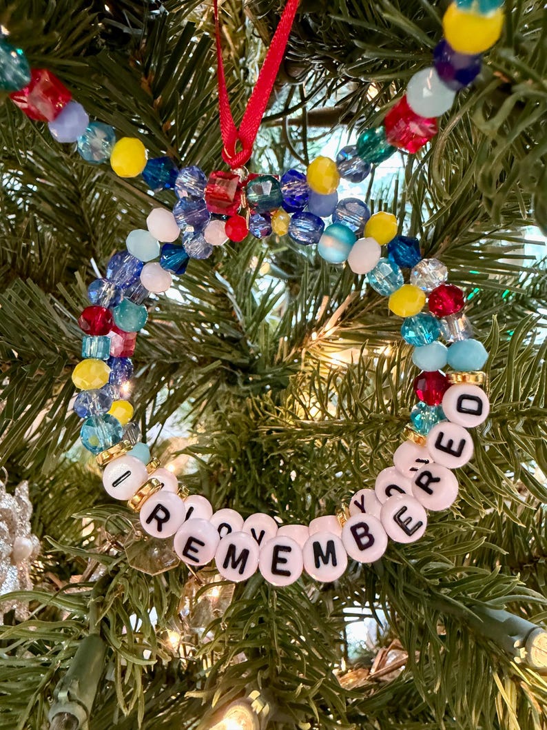May include: A circular ornament made of multi-coloured faceted beads in red, blue, yellow, and clear tones. The bottom section features white round beads with black letters spelling out I REMEMBERED. A red ribbon loop is attached to the top.