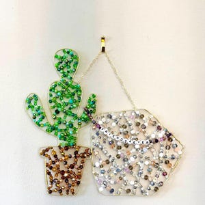 May include: A beaded cactus and a geometric shape with the text "FRIENDS 4 EVER" hang from a gold chain. The cactus is green and brown, while the other shape is filled with clear, pink, and purple beads.