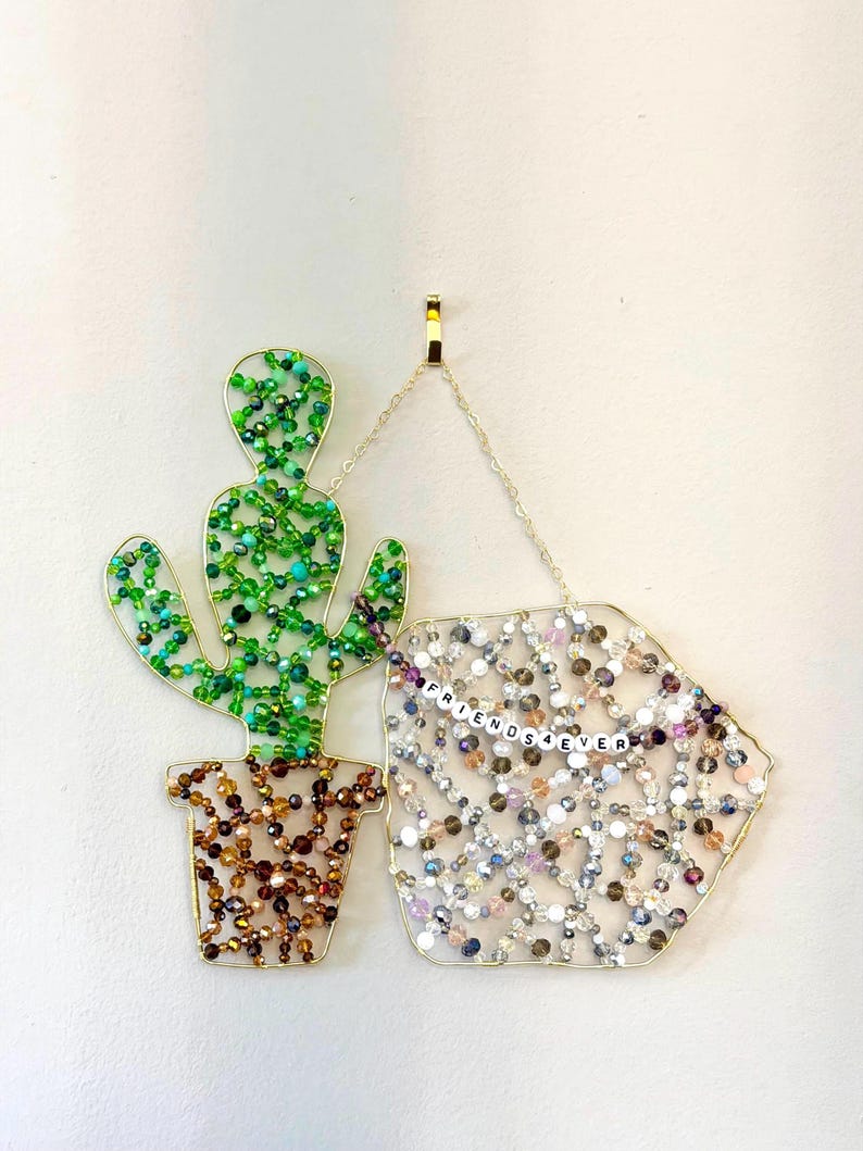 May include: A beaded cactus and a geometric shape with the text "FRIENDS 4 EVER" hang from a gold chain. The cactus is green and brown, while the other shape is filled with clear, pink, and purple beads.