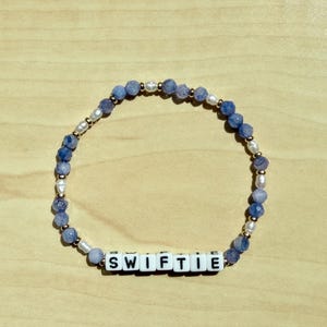May include: A beaded bracelet featuring blue and white beads, interspersed with pearls and gold-colored accents. The bracelet spells out "SWIFTIE" in white letter beads. The bracelet is displayed on a light brown wooden surface.