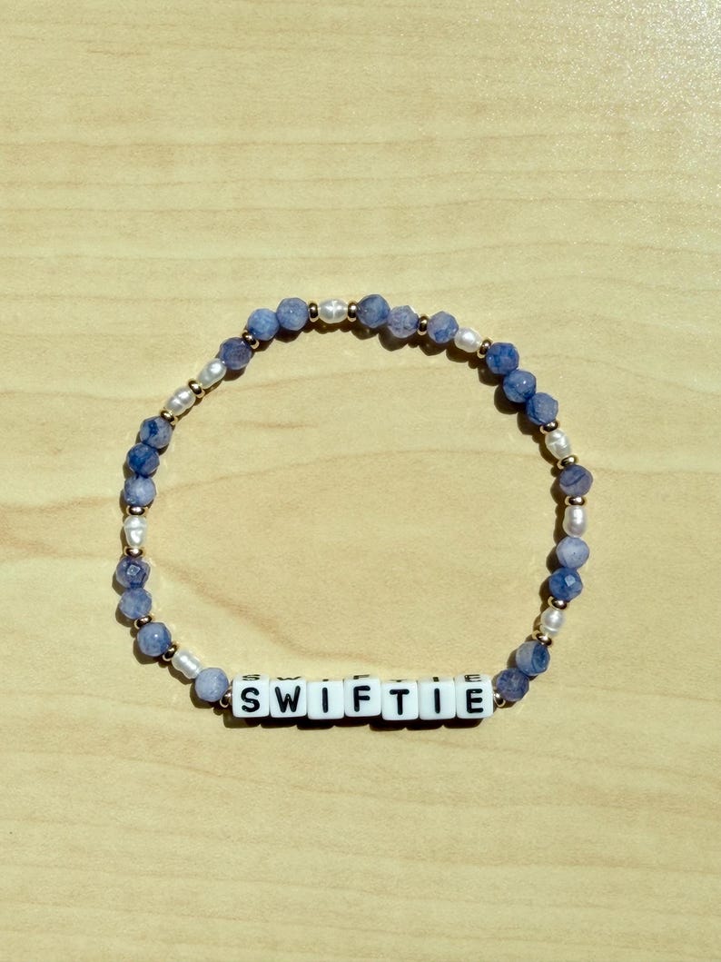 May include: A beaded bracelet featuring blue and white beads, interspersed with pearls and gold-colored accents. The bracelet spells out "SWIFTIE" in white letter beads. The bracelet is displayed on a light brown wooden surface.