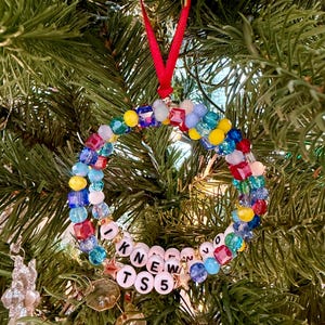K&ouml;nnte beinhalten: Ein kreisf&ouml;rmiger Weihnachtsschmuck aus bunten facettierten Perlen in Rot, Blau, Gelb und Hellviolett. Das Ornament enth&auml;lt wei&szlig;e Buchstabenperlen mit der Aufschrift I KNEW TS 5, akzentuiert durch kleine goldene Sternanh&auml;nger und eine rote Bandschlaufe.