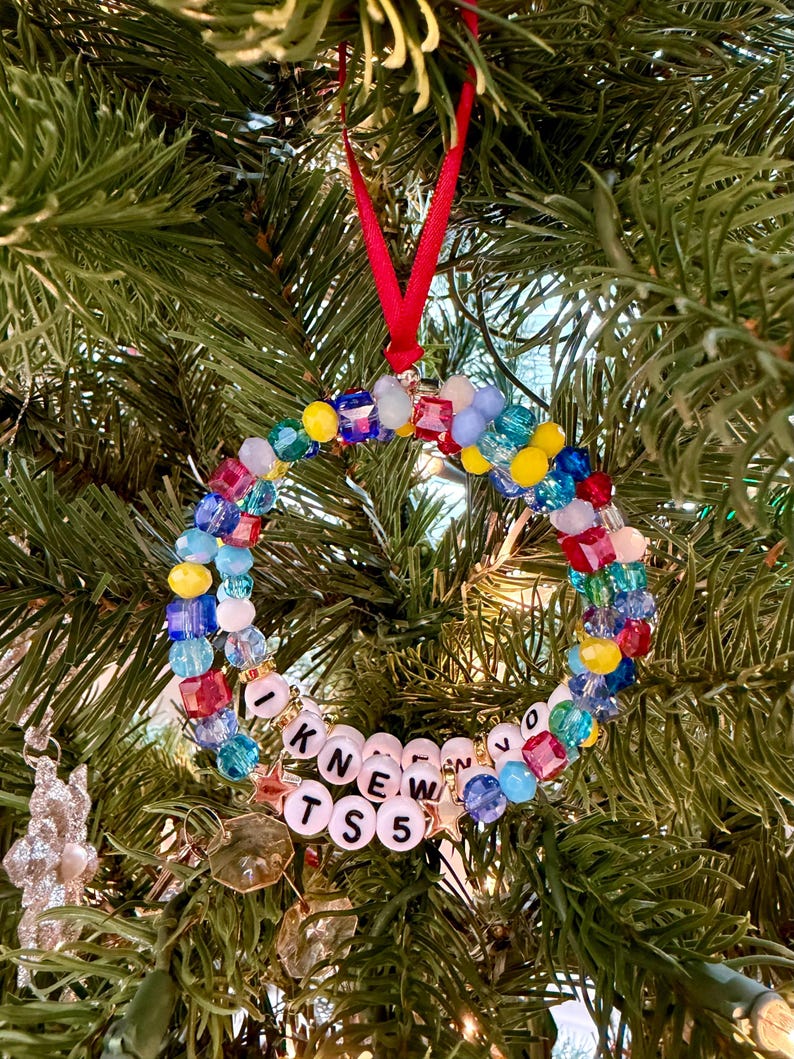 K&ouml;nnte beinhalten: Ein kreisf&ouml;rmiger Weihnachtsschmuck aus bunten facettierten Perlen in Rot, Blau, Gelb und Hellviolett. Das Ornament enth&auml;lt wei&szlig;e Buchstabenperlen mit der Aufschrift I KNEW TS 5, akzentuiert durch kleine goldene Sternanh&auml;nger und eine rote Bandschlaufe.