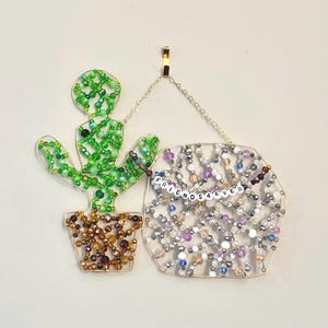 May include: A beaded cactus and sign with the words "FRIENDS 4 EVER". The cactus is green and brown, and the sign is filled with colorful beads. The artwork is hanging from a gold hook.
