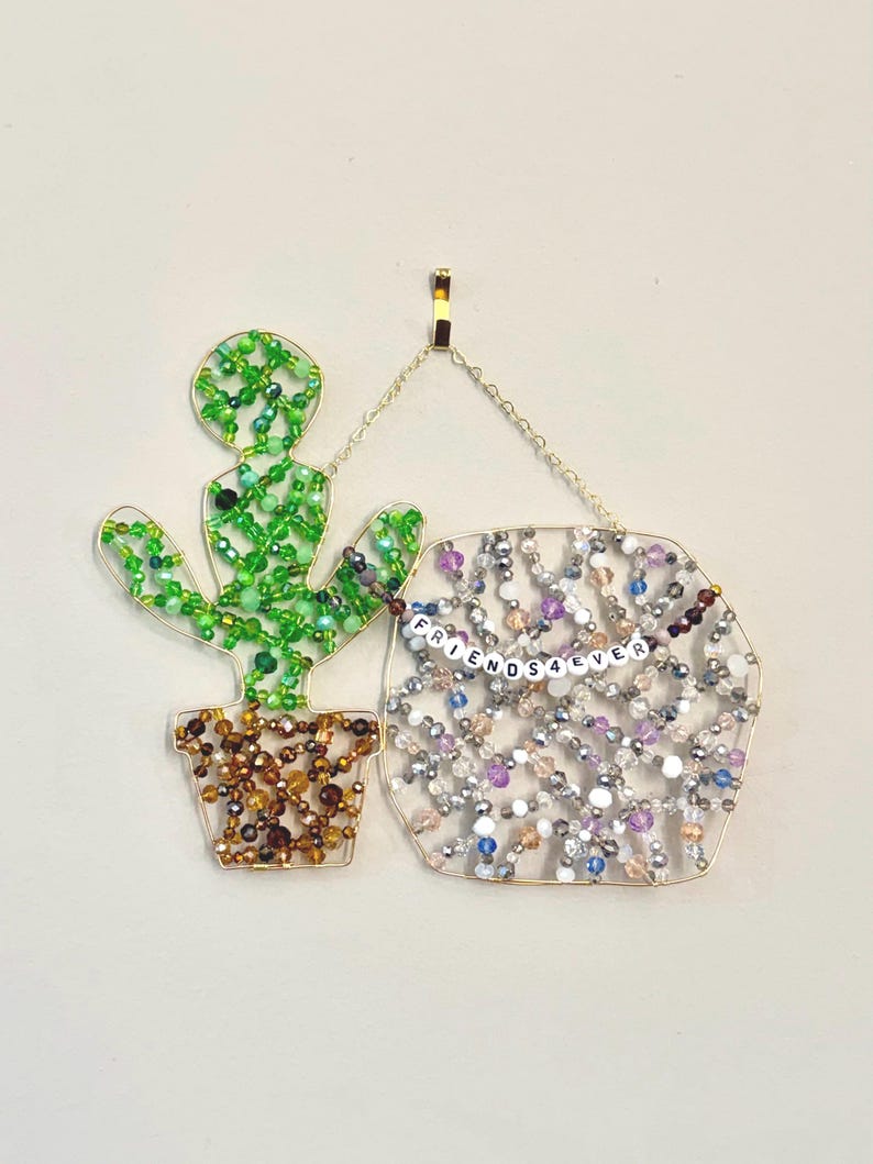 May include: A beaded cactus and sign with the words "FRIENDS 4 EVER". The cactus is green and brown, and the sign is filled with colorful beads. The artwork is hanging from a gold hook.