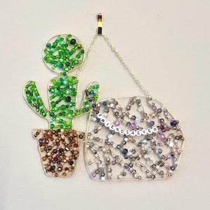 May include: A beaded cactus and a beaded sign that says "FRIENDS4EVER" hang from a gold-colored chain. The cactus is green and brown, and the sign is a mix of silver, purple, and clear beads.