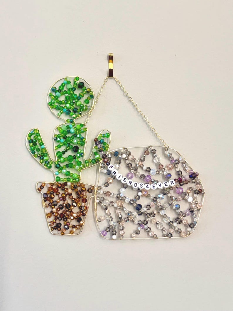 May include: A beaded cactus and a beaded sign that says "FRIENDS4EVER" hang from a gold-colored chain. The cactus is green and brown, and the sign is a mix of silver, purple, and clear beads.