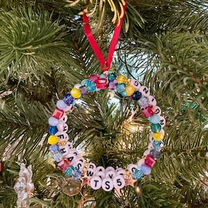 May include: A circular ornament made of multi-colored faceted beads and white letter beads spelling out "HANGING HAS DAYS TS 5". The ornament is suspended by a thin red ribbon and features small gold star accents interspersed among the beads.