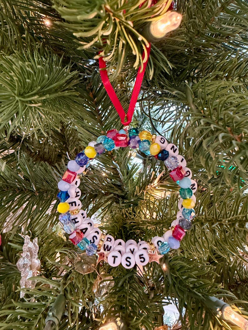 May include: A circular ornament made of multi-colored faceted beads and white letter beads spelling out "HANGING HAS DAYS TS 5". The ornament is suspended by a thin red ribbon and features small gold star accents interspersed among the beads.