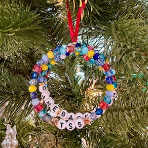 Puede incluir: Un adorno navide&ntilde;o circular hecho de una variedad de cuentas coloridas en tonos azul, amarillo y rojo, con cuentas de letras blancas que deletrean "CAME BACK TS 5" y un peque&ntilde;o dije en forma de estrella, suspendido por una cinta roja.