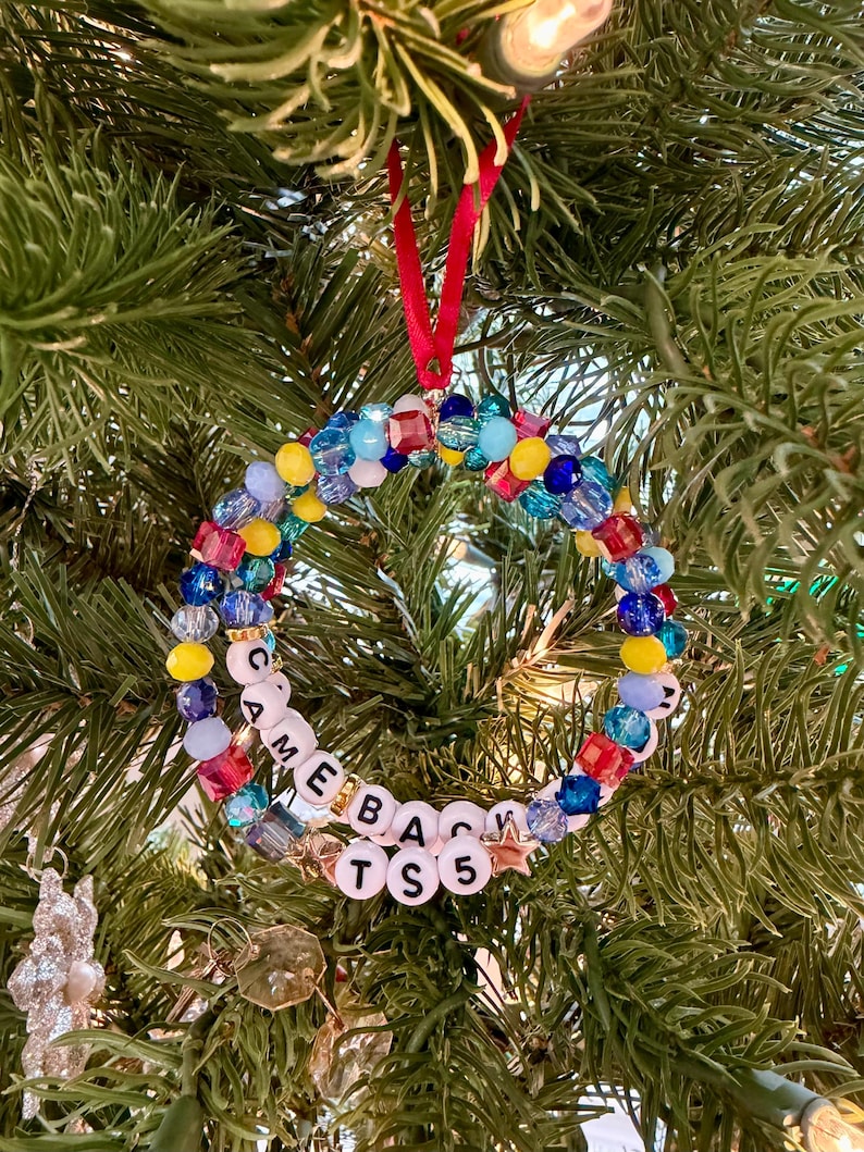 Puede incluir: Un adorno navide&ntilde;o circular hecho de una variedad de cuentas coloridas en tonos azul, amarillo y rojo, con cuentas de letras blancas que deletrean "CAME BACK TS 5" y un peque&ntilde;o dije en forma de estrella, suspendido por una cinta roja.