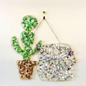 May include: A beaded wall hanging featuring a cactus and a sign that reads "FRIENDS 4 EVER." The cactus is green with a brown pot, and the sign is filled with clear and iridescent beads. The artwork is made with gold wire.