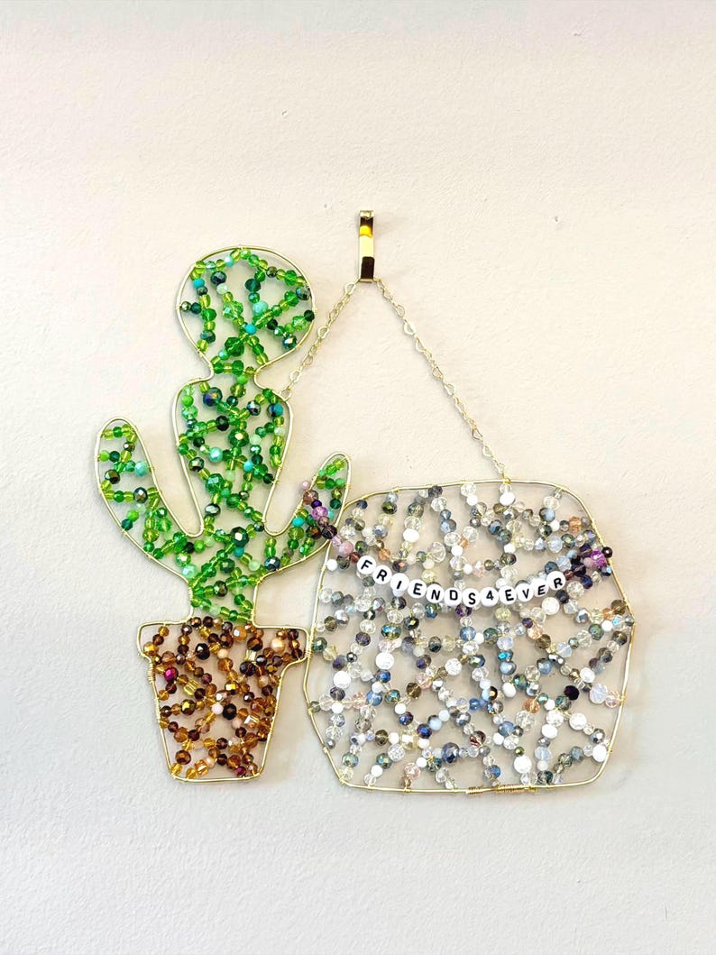 May include: A beaded wall hanging featuring a cactus and a sign that reads "FRIENDS 4 EVER." The cactus is green with a brown pot, and the sign is filled with clear and iridescent beads. The artwork is made with gold wire.