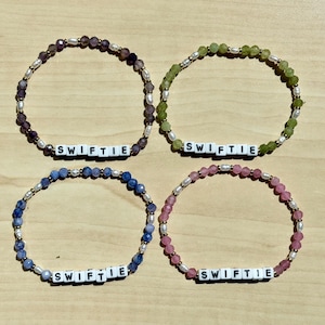 May include: Four beaded bracelets in shades of purple, green, blue, and pink. Each bracelet features the word "SWIFTIE" spelled out in white letter beads, interspersed with small, round beads and white pearls. The bracelets are displayed on a light wood surface.