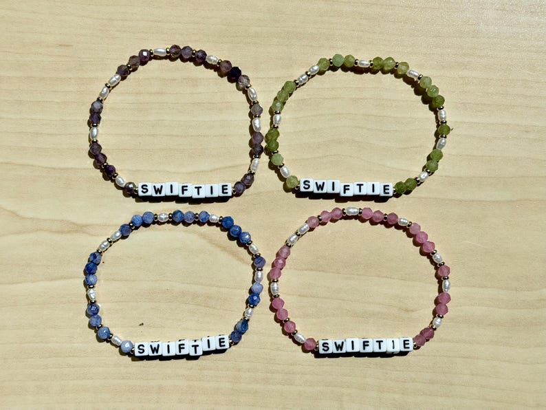May include: Four beaded bracelets in shades of purple, green, blue, and pink. Each bracelet features the word "SWIFTIE" spelled out in white letter beads, interspersed with small, round beads and white pearls. The bracelets are displayed on a light wood surface.