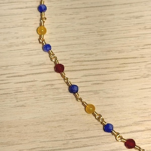 May include: A delicate gold-tone chain necklace featuring small, faceted, multi-coloured beads in shades of red, blue, and yellow. The beads are individually wire-wrapped and spaced evenly along the thin metallic chain, creating a repeating pattern.