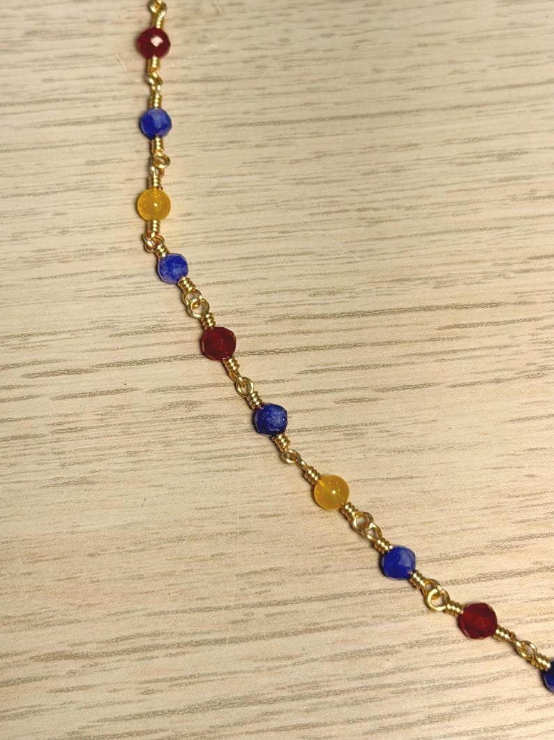 May include: A delicate gold-tone chain necklace featuring small, faceted, multi-coloured beads in shades of red, blue, and yellow. The beads are individually wire-wrapped and spaced evenly along the thin metallic chain, creating a repeating pattern.