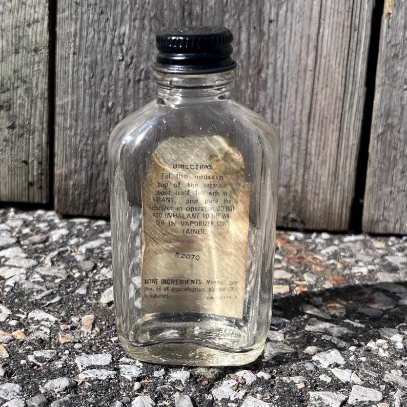 May include: A clear glass bottle with a black cap. The bottle has a label with text including "DIRECTIONS" and "ACTIVE INGREDIENTS". The bottle is rectangular with rounded edges. The label is tan and partially visible.