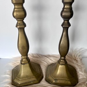 May include: Two antique brass candlesticks. Each candlestick has a hexagonal base, a detailed stem, and a flat top for holding a candle. The brass has a warm, aged patina. The candlesticks are displayed on a fluffy, light-colored surface.