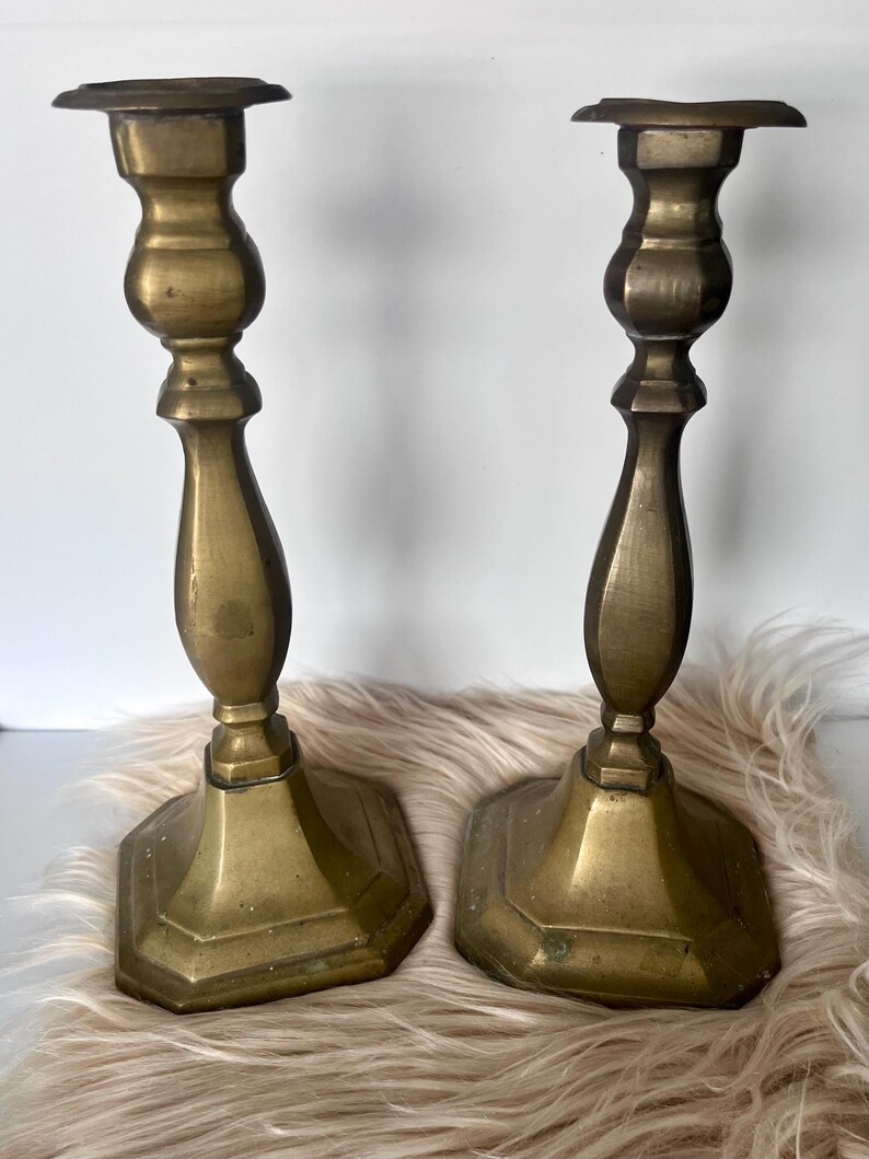 May include: Two antique brass candlesticks. Each candlestick has a hexagonal base, a detailed stem, and a flat top for holding a candle. The brass has a warm, aged patina. The candlesticks are displayed on a fluffy, light-colored surface.