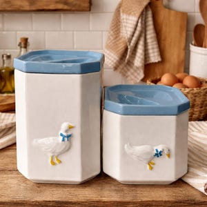Pode incluir: Dois potes de cozinha de cerâmica branca com tampas azuis. Cada pote apresenta um relevo de um ganso com um laço azul e pés amarelos. Os potes são hexagonais e estão sobre uma superfície de madeira.