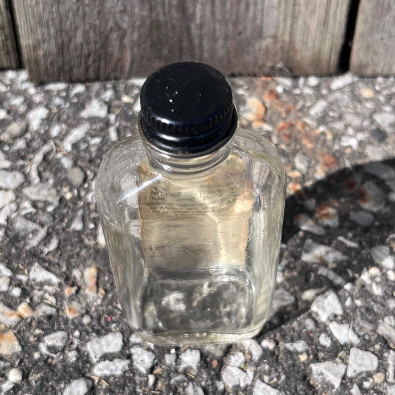 May include: A small, clear glass bottle with a black cap. The bottle has a square base and rounded shoulders. The cap is made of black plastic and screws onto the bottle. The bottle is sitting on a gray and white speckled surface.
