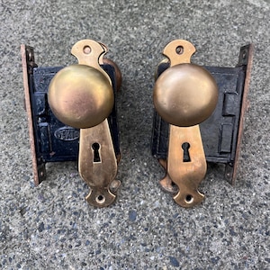 May include: Two matching door lock sets featuring round brass knobs and elongated brass backplates with keyholes. Each set includes a black cast iron rectangular lock box with a visible GENTO brand mark on the left unit's metal casing.