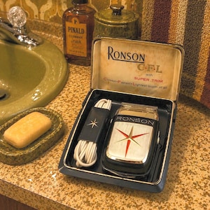 May include: Vintage Ronson CFL electric shaver in a black case. The shaver has a silver and black body with a red starburst design. The case also contains a black cord wrap with a star and a white cord. The text "RONSON CFL" is visible.