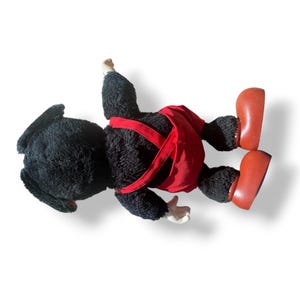 May include: A vintage plush toy monkey, lying on its back. The monkey has black fur, red overalls with suspenders, and large red shoes. Its hands and face are a lighter color.