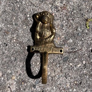 Puede incluir: Un gancho de pared de latón con un mono tapándose los oídos, con la inscripción "HEAR NO EVIL" debajo. El gancho es de color dorado cálido y está montado sobre una base rectangular.