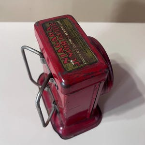 Puede incluir: Una linterna vintage roja de metal Niagara Junior Guide. La linterna tiene un cuerpo rectangular con una lente circular y un asa de metal. El texto "Niagara Junior Guide" es visible en la parte superior.