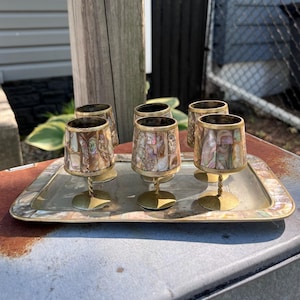 May include: A set of six small brass cups with iridescent abalone shell inlay, each featuring a twisted stem and a circular base, arranged on a matching rectangular brass tray with an abalone shell border and a smooth, reflective center surface.