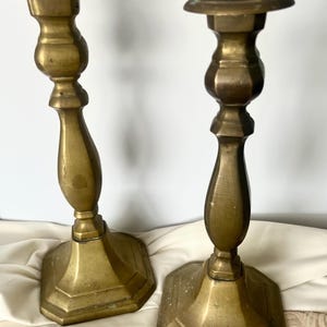 May include: Two antique brass candlesticks. Each candlestick has a hexagonal base, a decorative stem, and a flat top. The brass has a warm, aged patina, suggesting they are vintage pieces. The candlesticks are displayed on a white cloth.