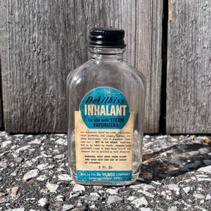 May include: A clear glass bottle with a black cap and a blue and white label that reads "DeVilbiss INHALANT" for use with steam vaporizers. The label also includes instructions and warnings. The bottle is 2 fl oz.