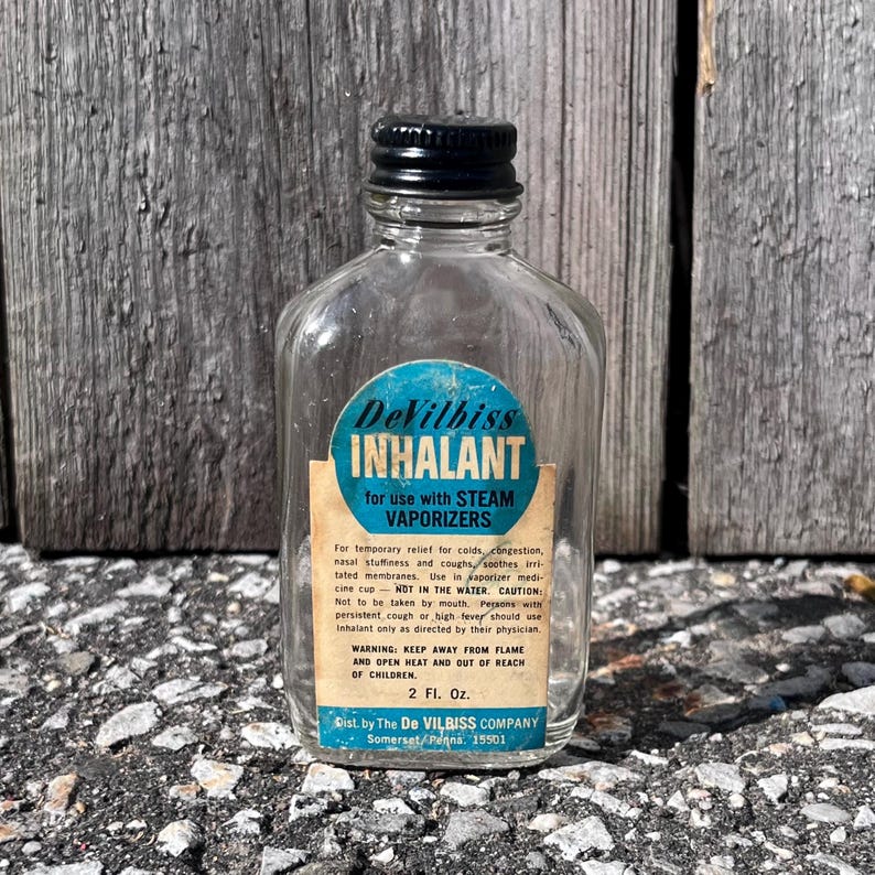 May include: A clear glass bottle with a black cap and a blue and white label that reads "DeVilbiss INHALANT" for use with steam vaporizers. The label also includes instructions and warnings. The bottle is 2 fl oz.