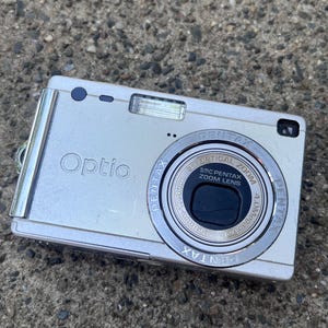 May include: A silver Pentax Optio digital camera featuring a 3x optical zoom lens with 4.0 megapixels. The front of the device displays the Optio logo, a flash unit, and a lens barrel with the text SMC Pentax Zoom Lens 5.8mm-17.4mm.